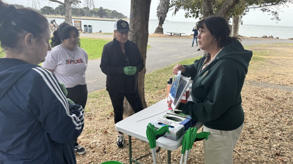 A group of BEI Construction volunteers gather around a table as a park staff member explains cleanup instructions, with grabbers and supplies laid out and the shoreline in the background.